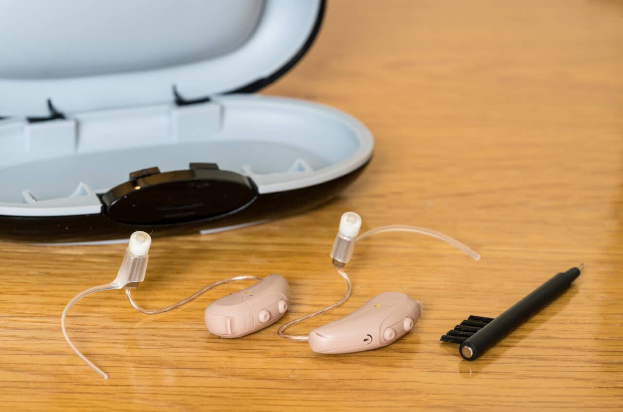 A pair of small hearing aids, case and a cleaning brush on a table.